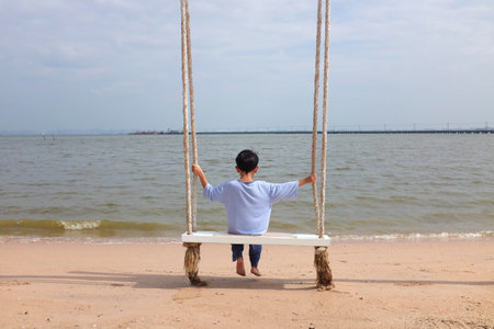 Asian boy child sitting on swing at the beach. Kid relax on summer holiday vacation.の写真素材
