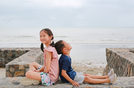 Portrait of cute Asian young girl child and little boy kid sitting together at seacoast. Sister and brother sit back against each other on cement wall.の写真素材