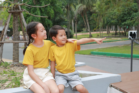 Portrait of Asian boy and girl are sitting in the park and pointing at something beside them,...