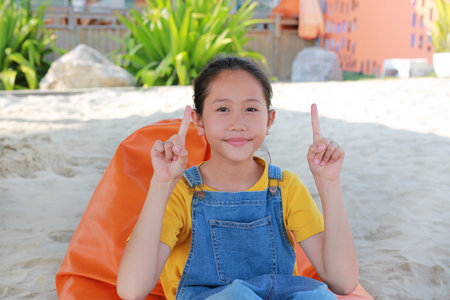 Closeup Asian girl in dungarees jean pointing up two forefinger while relax on orange sofa bed beach...