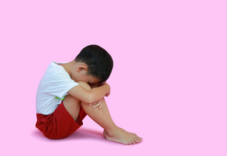 Unhappy Asian boy child age 7 years old sit hugging his knees and looking down isolated on pink studio background. Image full length with clipping path.の写真素材