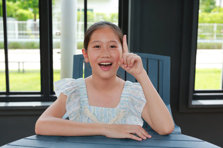 Cheerful Asian girl raising index finger while sitting on chair in pavilion.