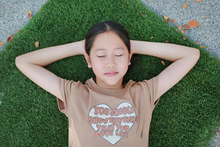Close-up top view of Asian girl child lying on green grass outdoors in summer park. Kid closed eyes...