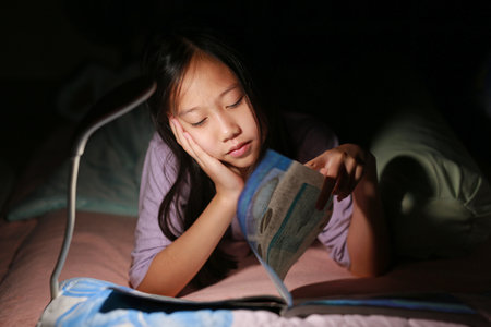 Closeup Asian girl kid reading a book under lamp light while lying on the bed at night. Soft focus.の写真素材