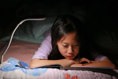 Closeup Asian girl kid reading a book under lamp light while lying on the bed at night. Soft focus.の写真素材