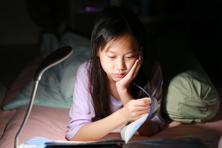 Portrait of Asian girl child reading a book with lamp light while lying on the bed at night. Close-up shot.の写真素材