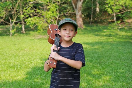 Asian boy holding ukulele on shoulder with looking camera while standing outdoor in the summer...