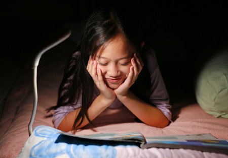 Cheerful Asian girl preteen reading a book under lamp light while lying on the bed at night. Close...