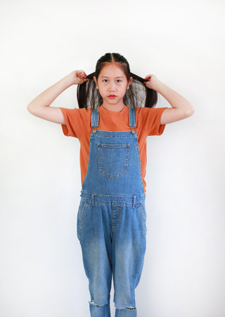 Asian girl in denim overalls tying her hair back to a ponytail standing against a white background.