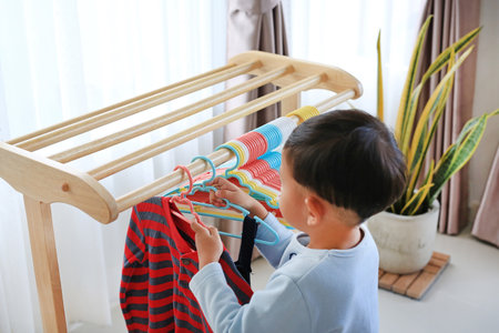 Little Asian boy using hanger hang up a clothes on wood rack.