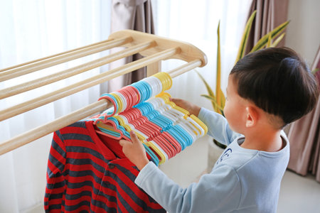 Asian little boy use hanger hang up a clothes on wood rack.