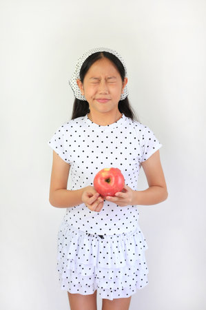 Portrait of  Asian girl in a polka dot outfit holds a red apple close to her chest with a scrunched, eyes-closed expression.の写真素材
