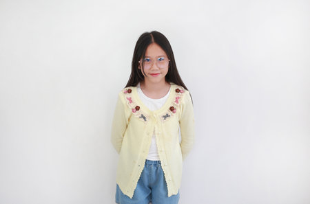 Cheerful young Asian girl wearing a pastel yellow cardigan with floral embroidery and glasses smiles...