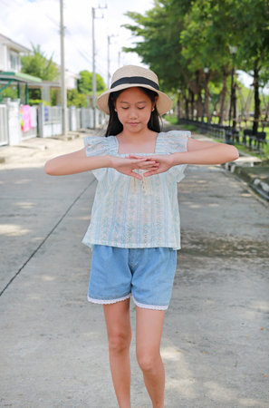 Calm young Asian girl wearing straw hat, summer top, and denim shorts is standing on a paved road outdoors, holding her hands together in front of her chest in focused gesture.の写真素材