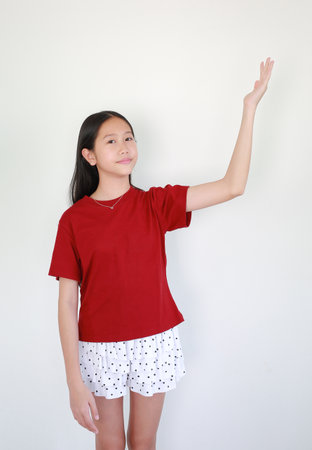 Portrait young Asian girl in red t-shirt and polka dot skirt standing against white background, smiling and raising her hand to point or present something.の写真素材