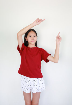 Asian girl in red t-shirt and polka dot skirt standing against white background, smiling happily while pointing her fingers upward to present a product or a creative idea.の写真素材