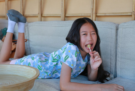 Portrait of charming young Asian girl lying down on a cozy sofa while enjoying a colorful lollipop.