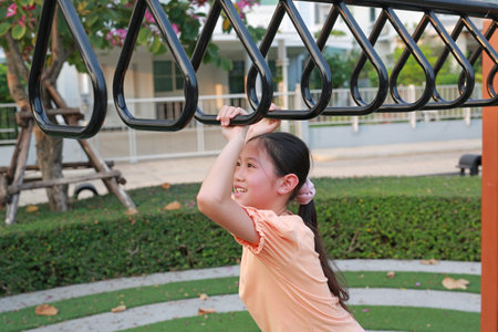 Cheerful young girl having fun and exercising on the overhead hanging rings at a playground during...