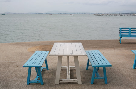 Serene coastal spot featuring a white wooden picnic table and blue benches, perfectly inviting someone to sit back, relax, and soak in the tranquil ocean view on a calm, overcast day.の写真素材