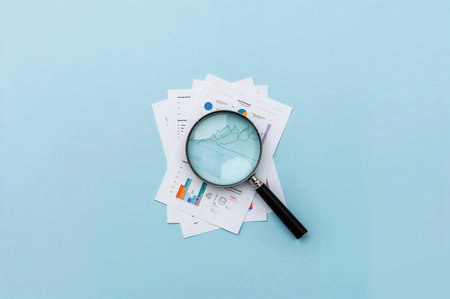 Magnifying glass remaining on data sheets with colorful charts. Business analytics or financial research is visualized on a light blue backdrop.の素材