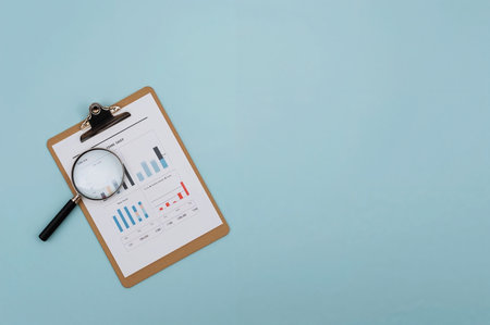 Clipboard with charts and a magnifying glass on a blue background. Examining data and financial reporting. Detailed financial analysis is shown.の素材
