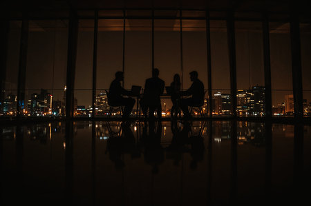 Silhouetted figures meet against a backdrop of city lights, reflecting on polished floors. Team discusses strategies, success, and collaboration.の素材