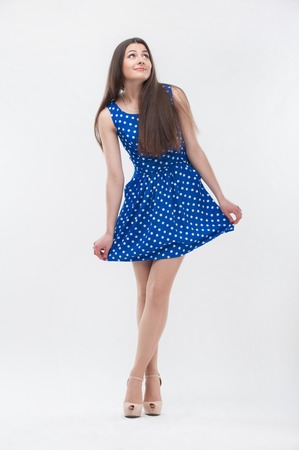 Emotional attractive young shy brunette woman with long hair in blue dress standing in full length isolated on white backgroundの写真素材