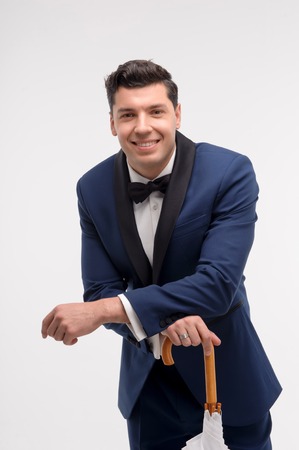 Fashion man wearing blue suit and black bow tie. Holding a stick umbrella. Studio shot against white wallの写真素材