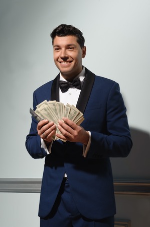 Happy handsome young brunet man holding a lot of money looking sideways while standing on blue wall backgroundの写真素材