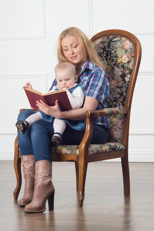 Happy attractive blonde mother sitting and reading a book with baby girl in arm chairの写真素材