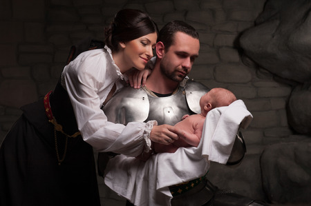 Portrait of a beautiful medieval couple, kneeling knight holding newborn baby and lady bending over them on dark stone backgroundの写真素材