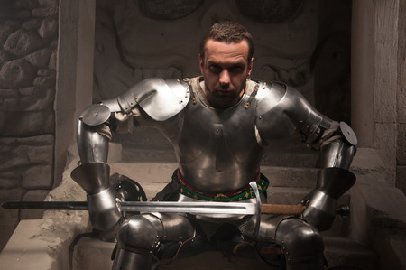 Closeup portrait of medieval knight in armor sitting on steps of ancient temple in smoke, dark backgroundの写真素材
