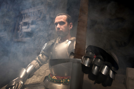 Waistup portrait of medieval knight in armor with sword sitting on steps of ancient temple in smoke, dark backgroundの写真素材