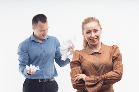 Portrait of young attractive cheerful employee smiling while her colleague on the background crumpling paper in anger, isolated on whiteの写真素材