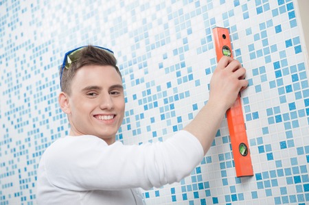 Young caucasian handsome worker using a spirit level on blue tile wall, interior background