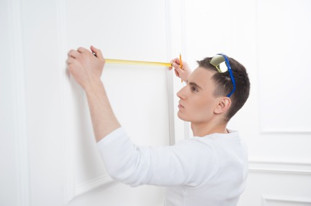 Young caucasian handsome smiling man in protective glasses with pencil using a measuring tape near...
