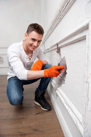 Repair, renovation and home concept  Young handsome man in sneakers aligns the white cracked wall with spatula smiling in cameraの写真素材
