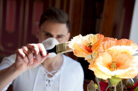 Repair, renovation and creative concept  Closeup portrait of young handsome worker in mask painting real bright flowers with paintbrush, interior shotの写真素材