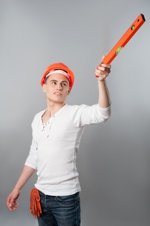 Portrait of a young smiling handsome worker in helmet showing something with spirit level isolated on grey backgroundの写真素材
