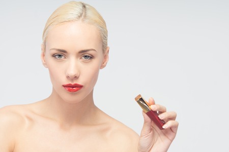 Portrait of sexy young woman looking at camera holding red lip gloss bottle, isolated on whiteの写真素材