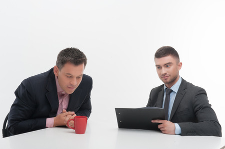 Senior and junior business people in elegant suits sitting at desk, boss looking in red cup,...