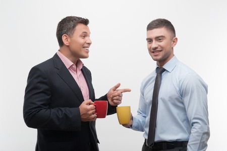 Senior and junior handsome smiling business people discuss something during coffee break holding red and yellow cups, isolated on white backgroundの写真素材