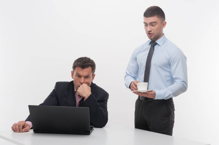 Young handsome office worker assistant brings coffee cup to senior manager sitting at desk with laptop isolated on whiteの写真素材