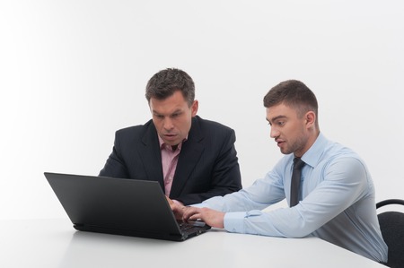 Senior and junior handsome business people discuss something sitting at desk, employee showing something on laptop to his boss, isolated on whiteの写真素材
