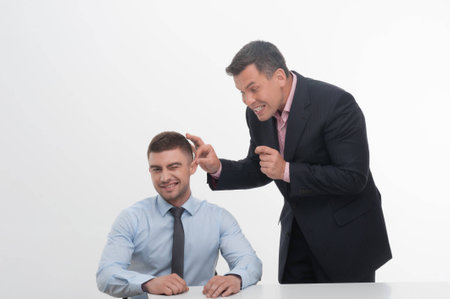 Senior mature manager chief standing by desk pulling his young junior employee ear, isolated on whiteの写真素材