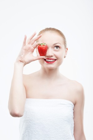 Waistup portrait of a young attractive smiling woman having fun with strawberry, covering her eye, isolated on whiteの写真素材