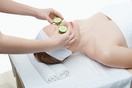 Beautiful young woman receiving facial mask of cucumber in beauty salon, hands of cosmetologist, isolated on whiteの写真素材