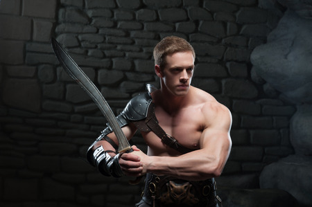 Half length portrait of young attractive warrior gladiator with muscular body posing with sword on dark background  Concept of masculine power, strengthの写真素材