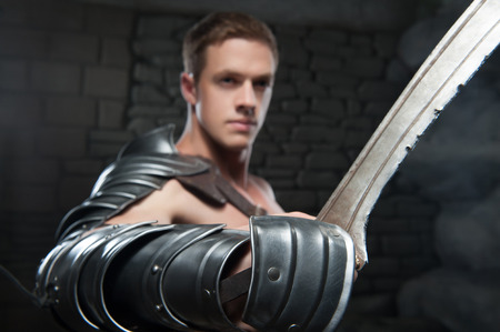 Closeup portrait of young attractive warrior gladiator with muscular body posing with sword on dark background  Selective focus, Concept of masculine power, strengthの写真素材