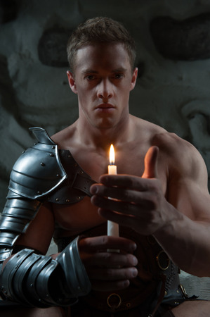 Waistup portrait of young handsome muscular man Gladiator in armour sitting on steps of ancient temple holding candle against dark backgroundの写真素材